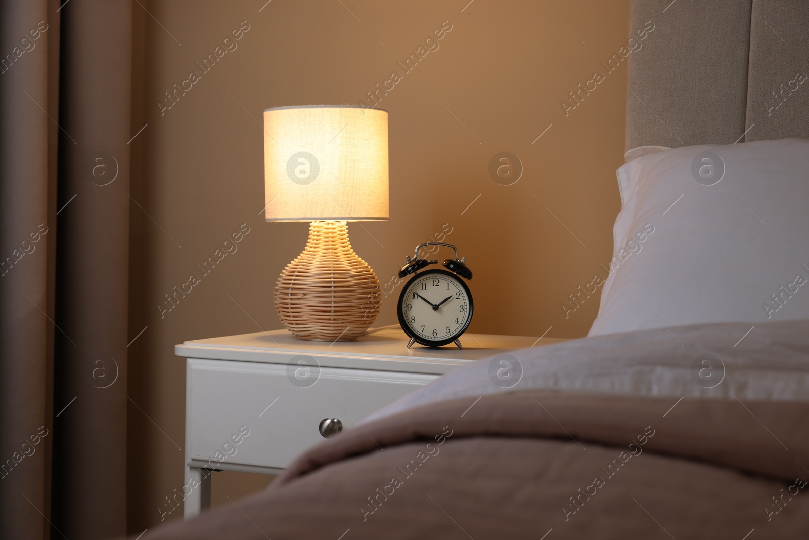 Photo of Glowing lamp and alarm clock on nightstand in bedroom