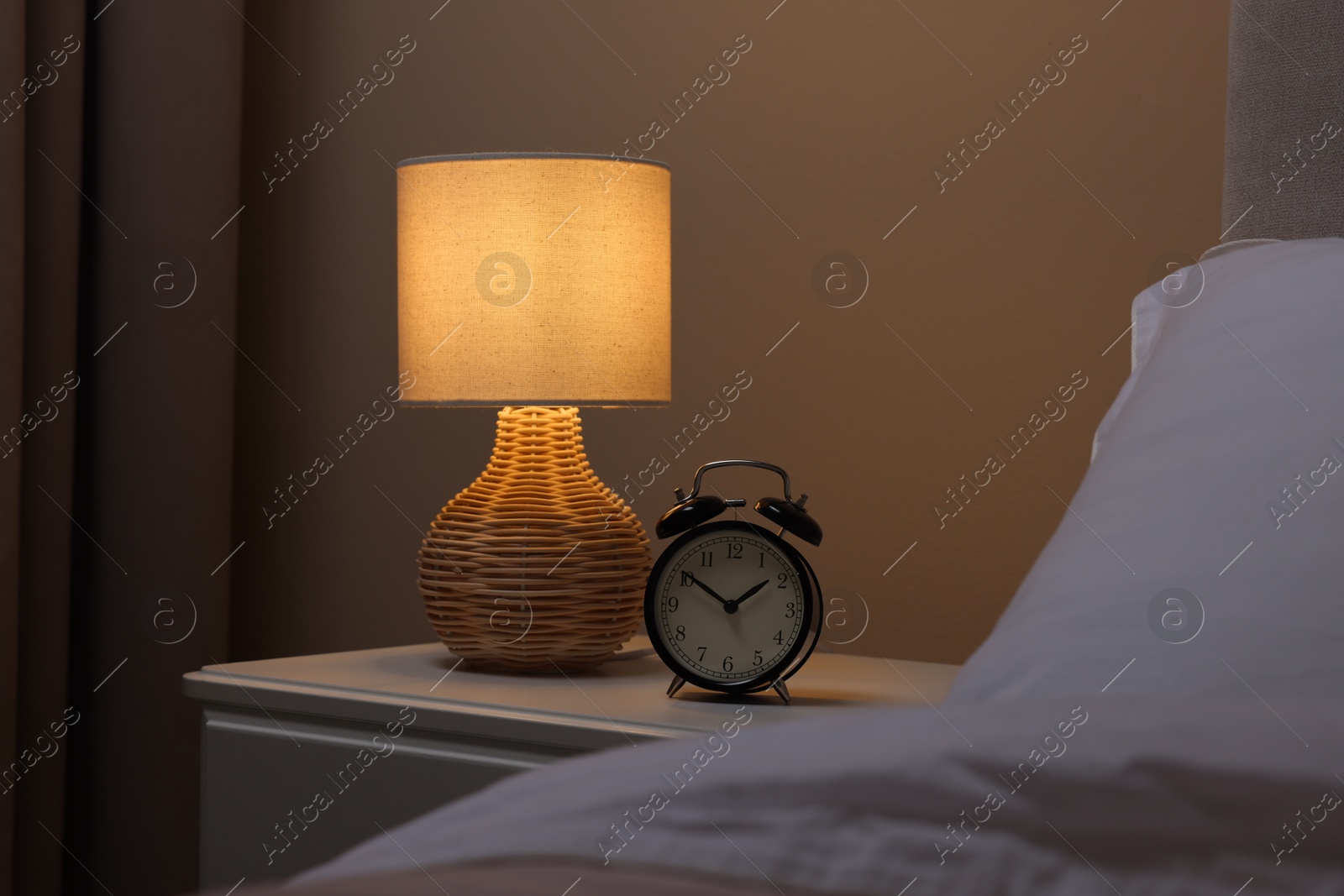 Photo of Glowing lamp and alarm clock on nightstand in bedroom