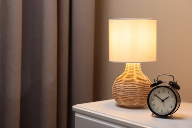 Photo of Glowing lamp and alarm clock on nightstand in bedroom. Space for text