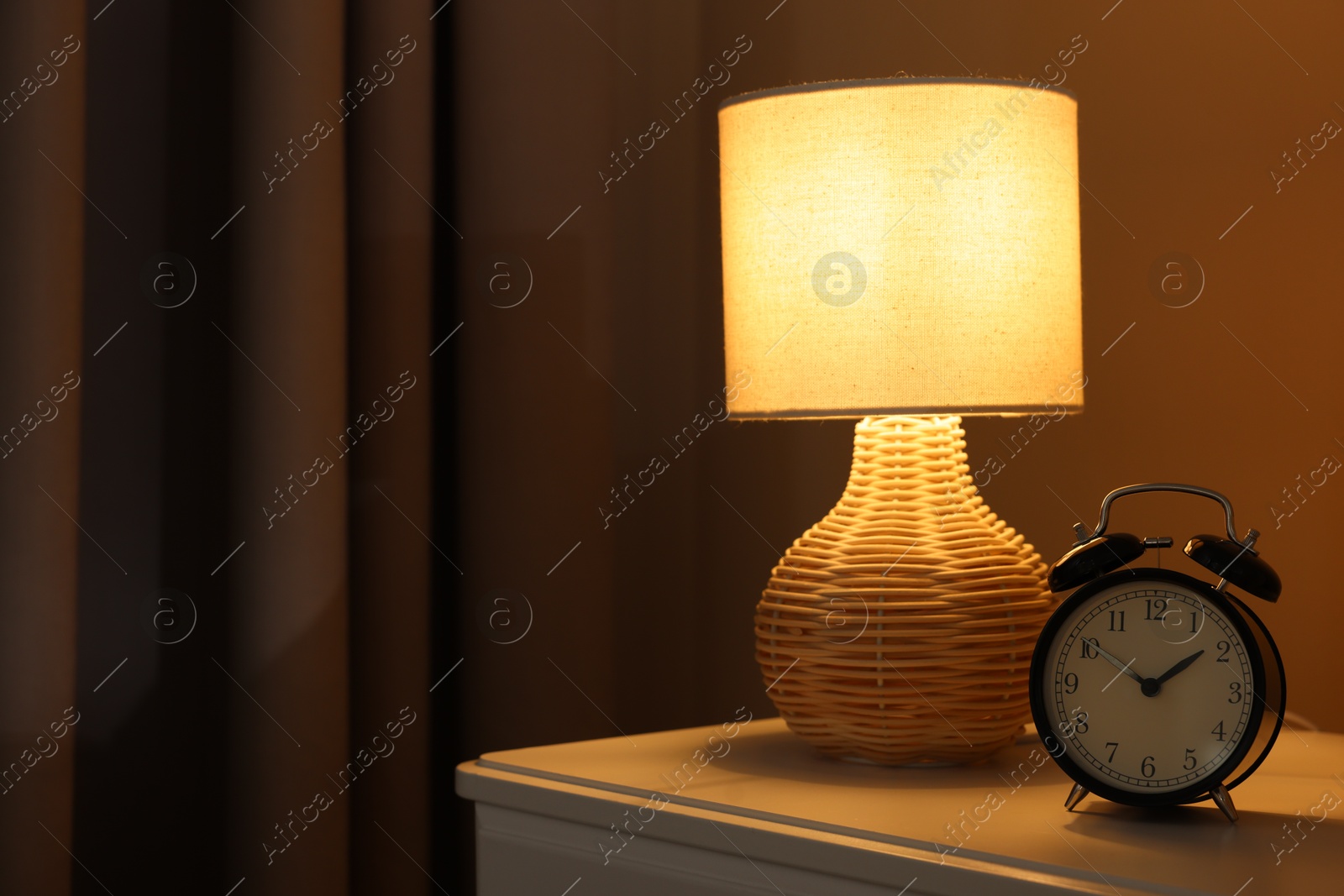 Photo of Glowing lamp and alarm clock on nightstand in bedroom. Space for text