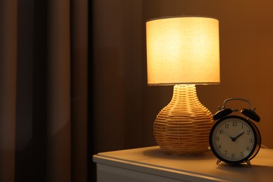 Photo of Glowing lamp and alarm clock on nightstand in bedroom. Space for text