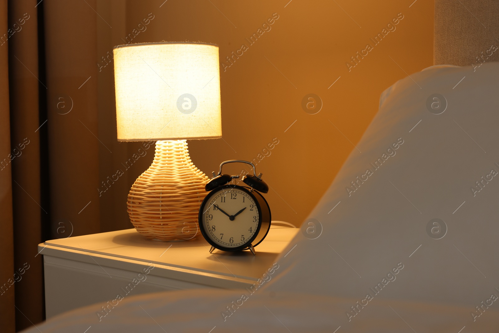 Photo of Glowing lamp and alarm clock on nightstand in bedroom