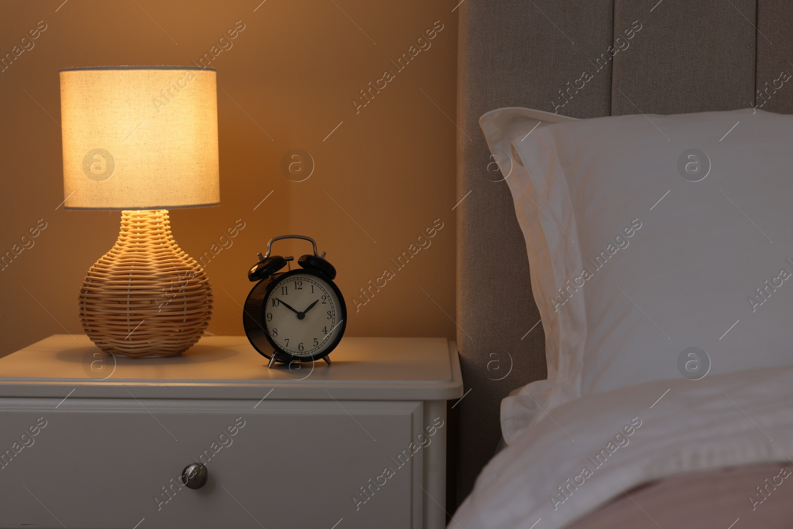 Photo of Glowing lamp and alarm clock on nightstand in bedroom