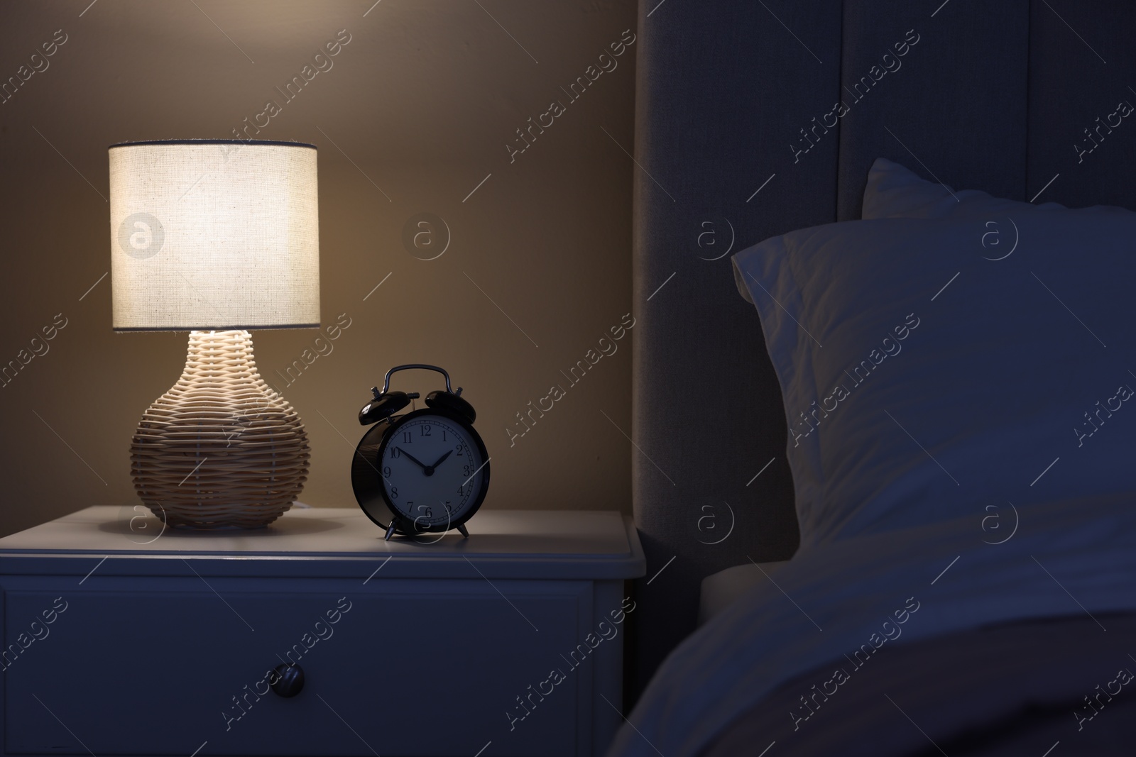 Photo of Glowing lamp and alarm clock on nightstand in bedroom