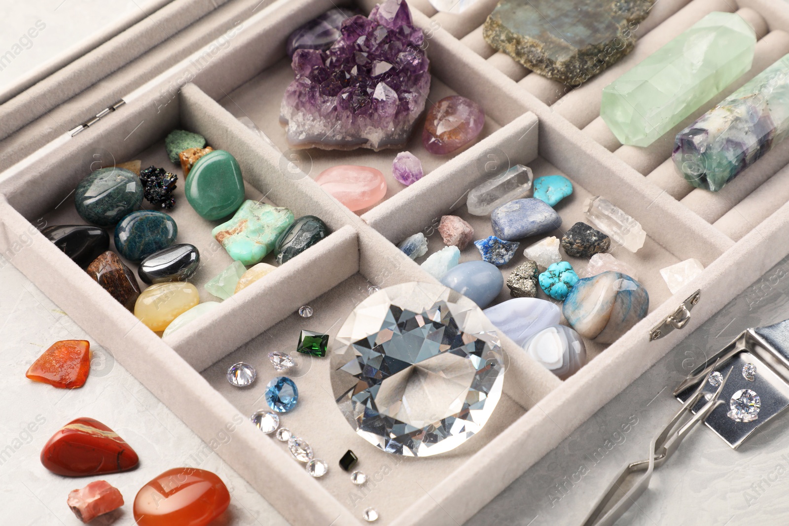 Photo of Box with beautiful gemstones and jeweler's tools on light table, closeup