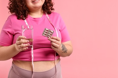 Photo of Happy plus size woman holding blister of weight loss supplements and glass of water on pink background, closeup. Space for text