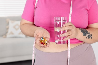 Photo of Plus size woman holding pile of weight loss supplements and glass with water at home, closeup