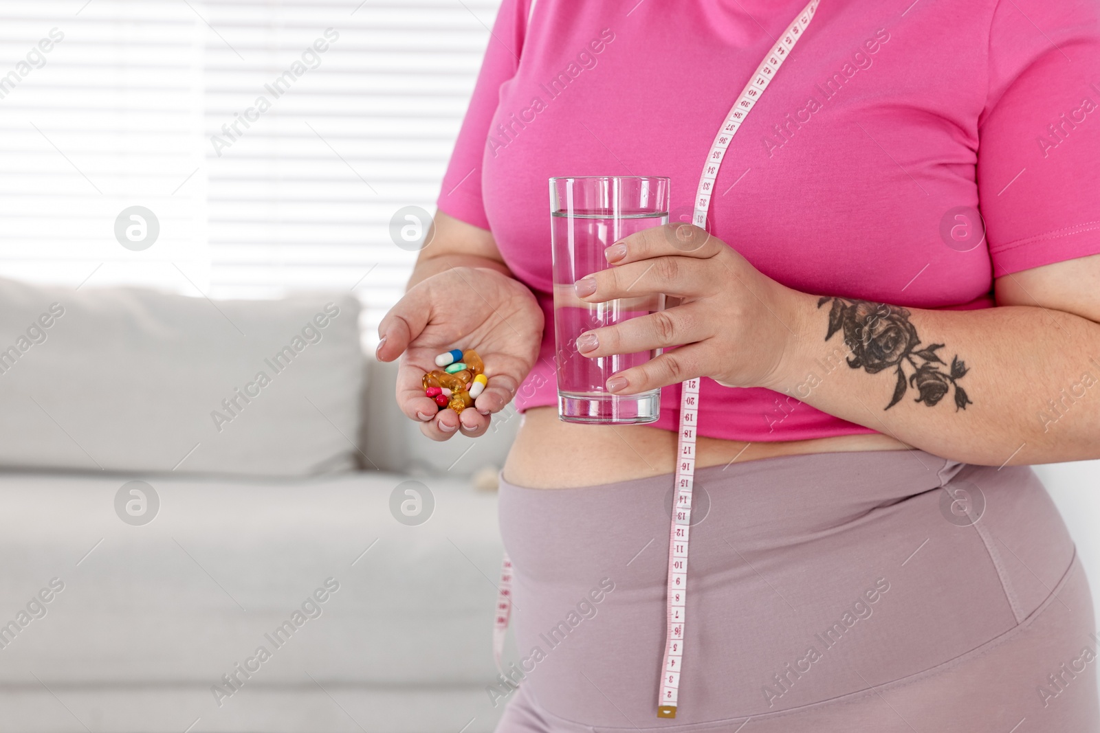 Photo of Plus size woman holding pile of weight loss supplements and glass with water at home, closeup. Space for text