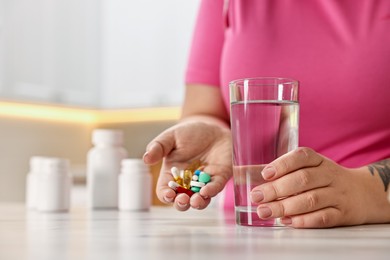 Photo of Plus size woman holding pile of weight loss supplements and glass with water at marble countertop in kitchen, closeup. Space for text