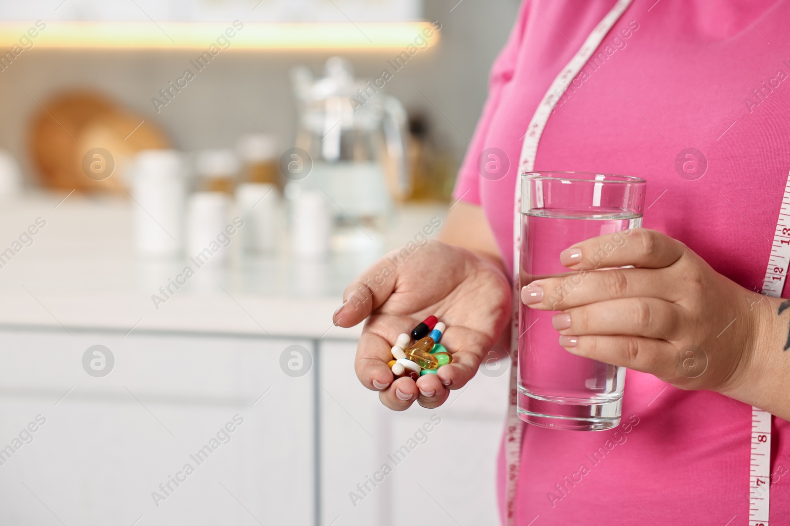 Photo of Plus size woman holding pile of weight loss supplements and glass with water in kitchen, closeup. Space for text