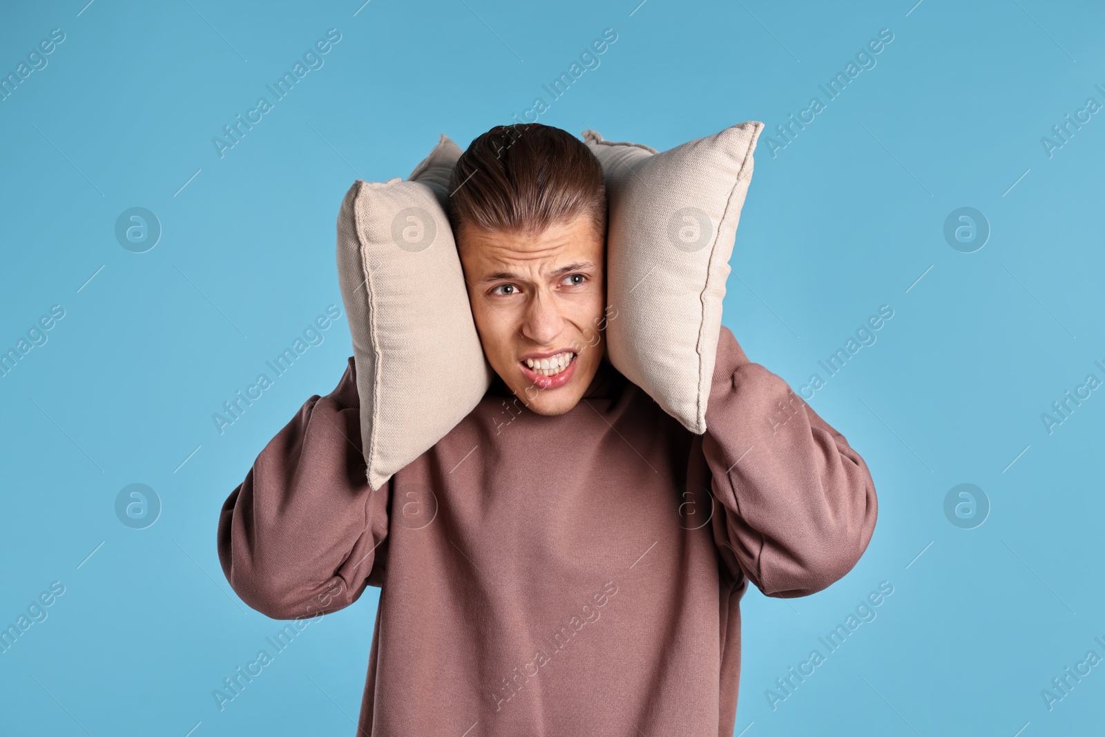 Photo of Annoyed young man covering his ears with pillows due to loud sound on light blue background