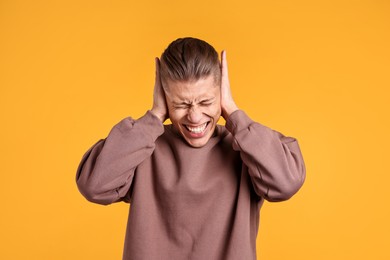 Photo of Annoyed young man covering his ears due to loud sound on orange background