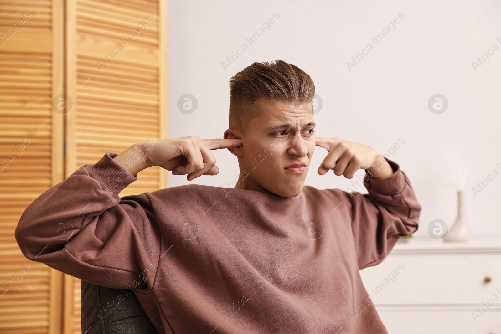 Photo of Annoyed young man covering his ears due to loud sound at home