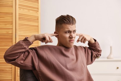 Photo of Annoyed young man covering his ears due to loud sound at home