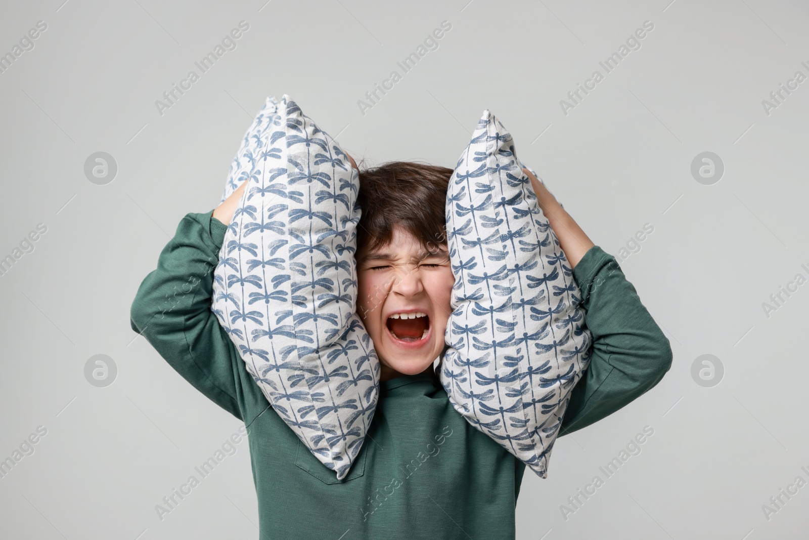 Photo of Annoyed boy covering his ears with pillows due to loud sound on grey background