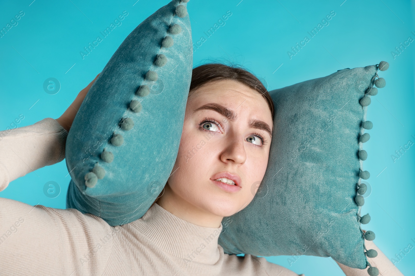 Photo of Distressed woman covering her ears with pillows from loud noise on blue background