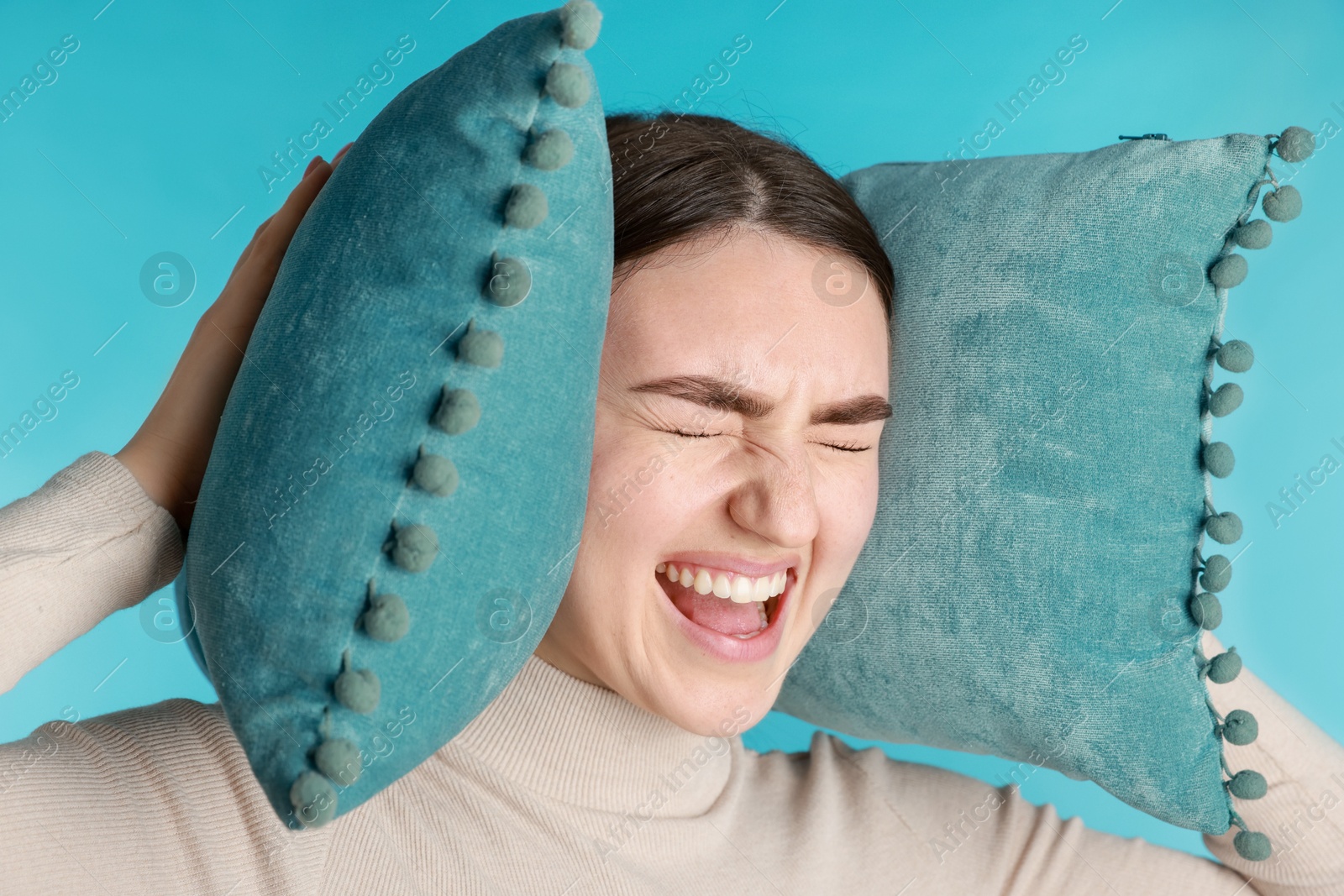 Photo of Frustrated woman covering her ears with pillows from loud noise on blue background