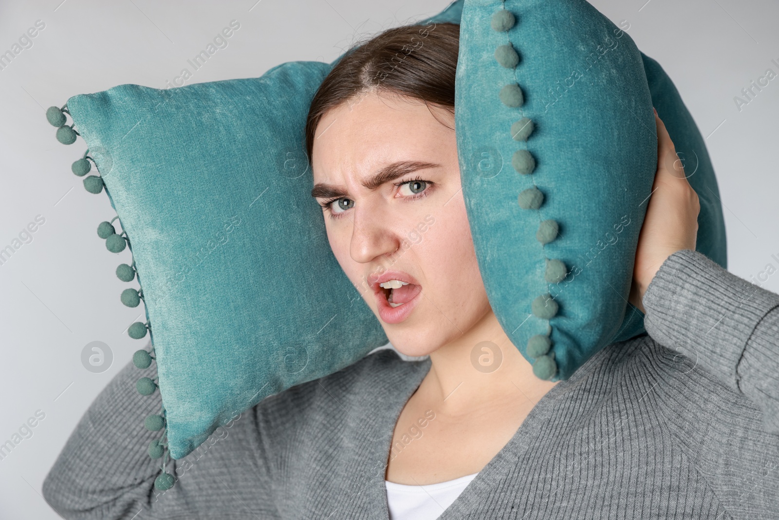 Photo of Frustrated woman covering her ears with pillows from loud noise on light grey background