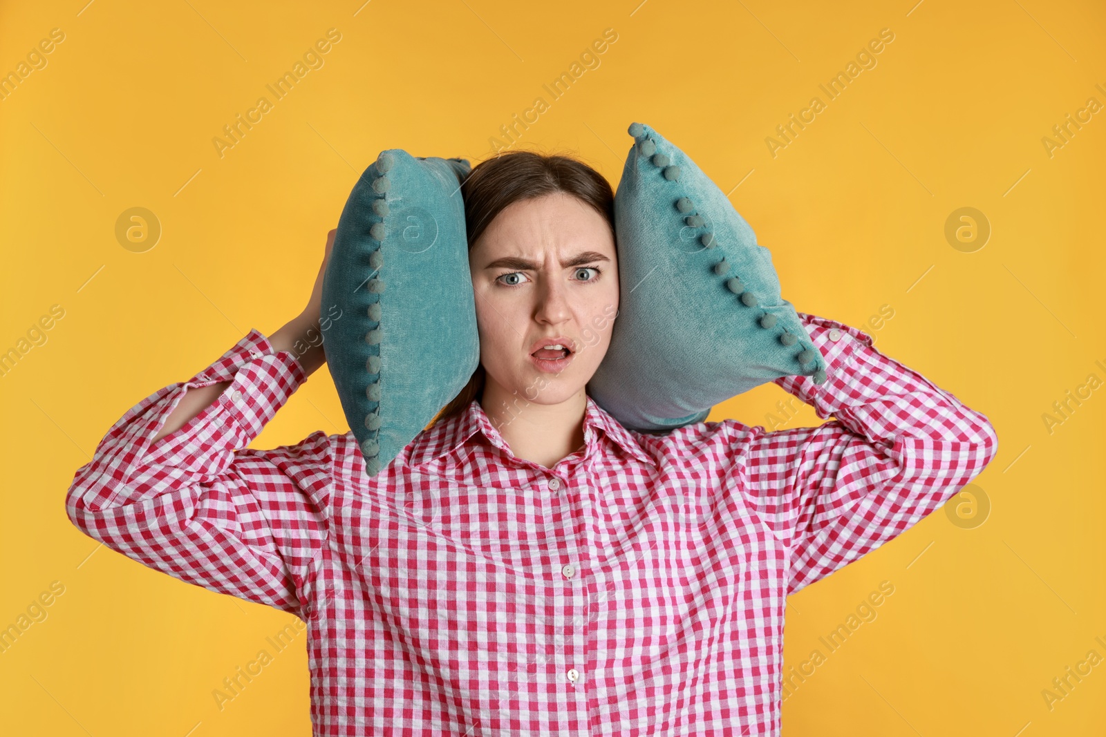 Photo of Distressed woman covering her ears with pillows from loud noise on yellow background