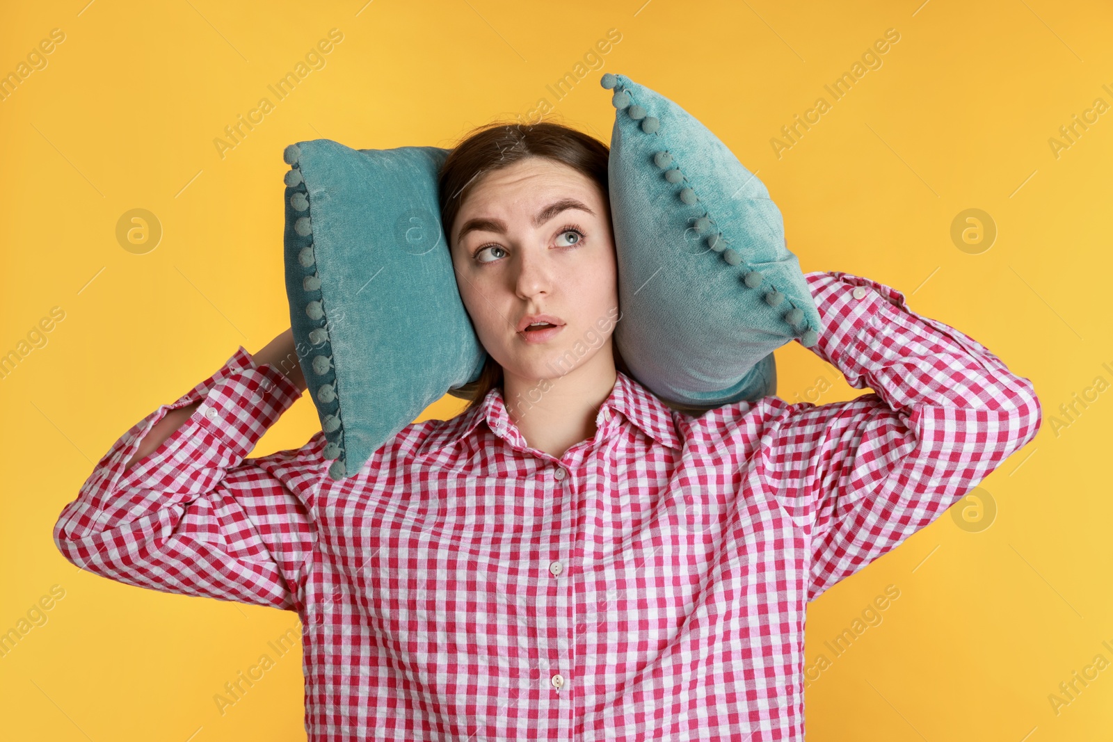 Photo of Distressed woman covering her ears with pillows from loud noise on yellow background