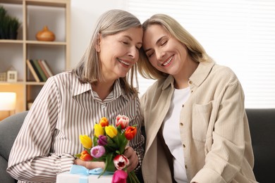 Photo of Smiling woman with bouquet of tulips, gift and her daughter on sofa at home. Happy Mother's Day