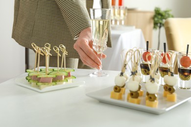 Photo of Many different tasty canapes on white table. Woman with glass of wine indoors, closeup