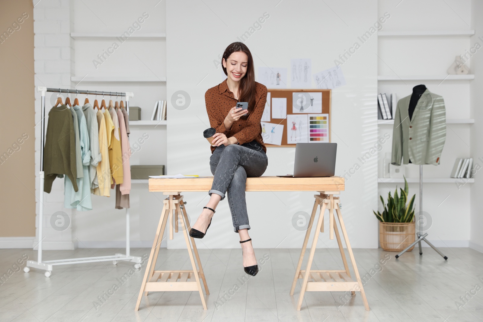 Photo of Fashion designer with paper cup of coffee using smartphone in workshop