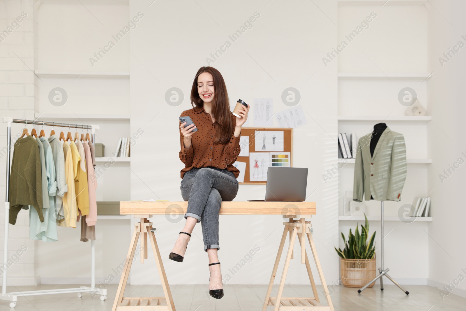 Photo of Fashion designer with paper cup of coffee using smartphone in workshop