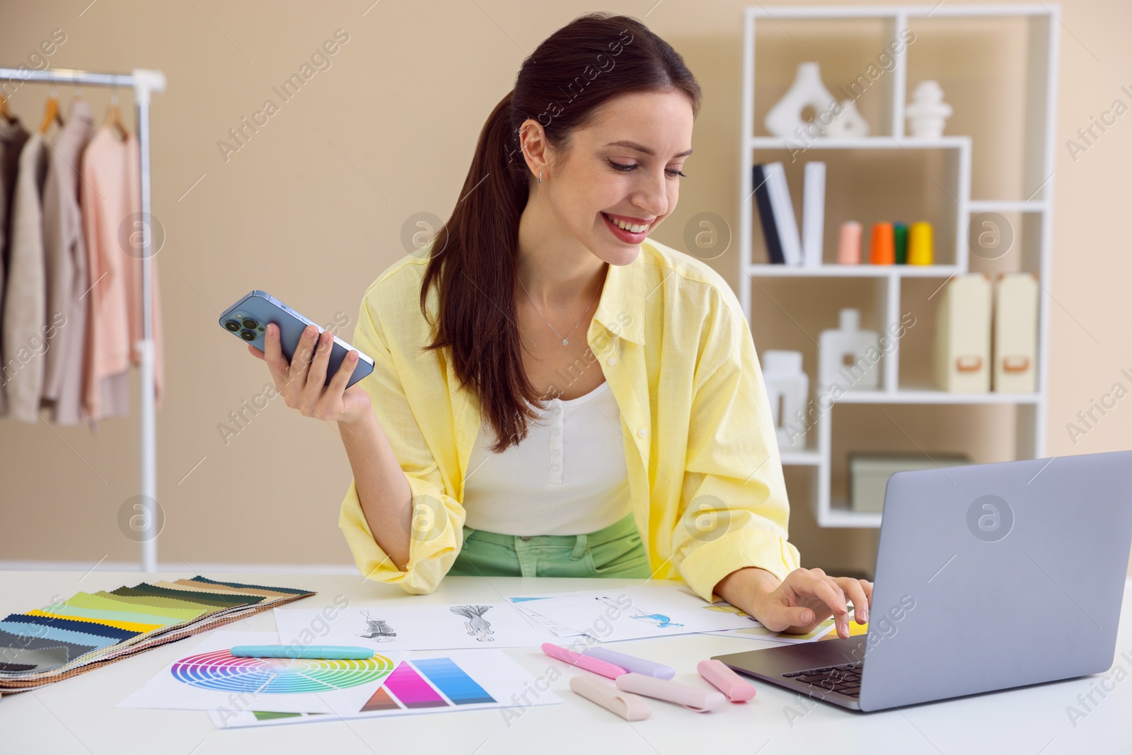 Photo of Fashion designer with smartphone working on laptop at white table in workshop