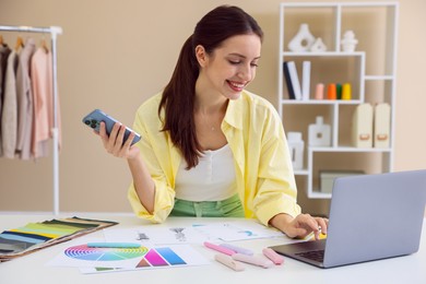 Photo of Fashion designer with smartphone working on laptop at white table in workshop