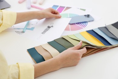 Photo of Fashion designer with sketch of outfit choosing fabric among colorful samples at white table in workshop, closeup