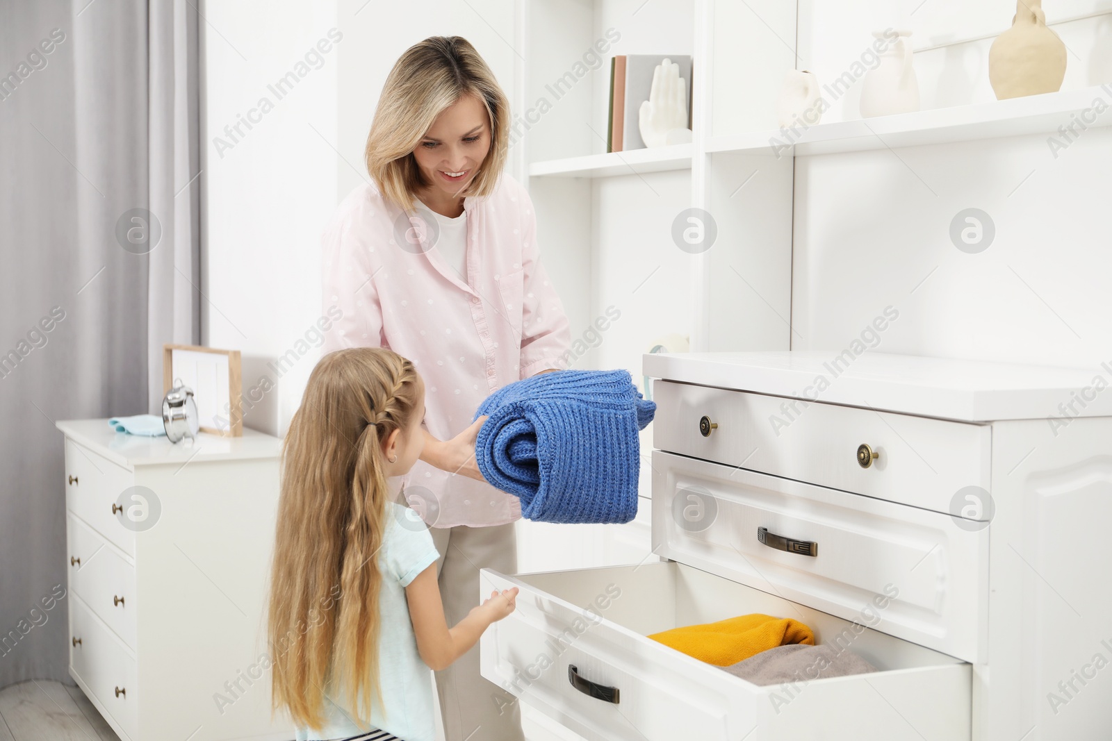 Photo of Little girl helping her mother putting clothes into drawers at home