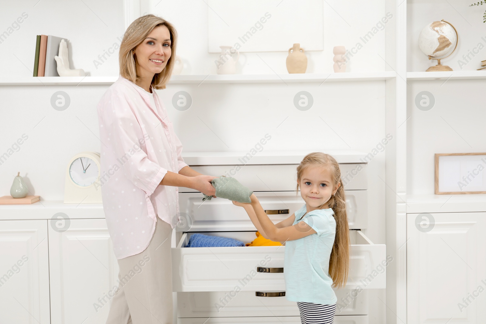 Photo of Little girl helping her mother putting clothes into drawers at home