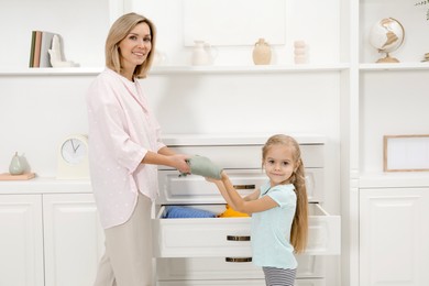 Photo of Little girl helping her mother putting clothes into drawers at home