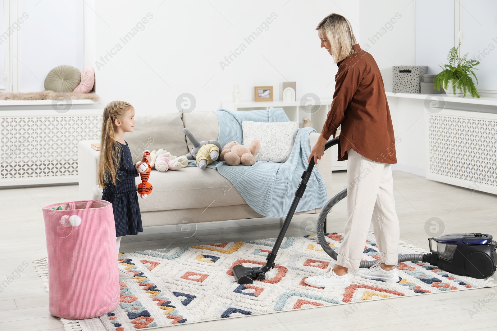 Photo of Little helper. Girl putting away toys while her mother vacuuming at home