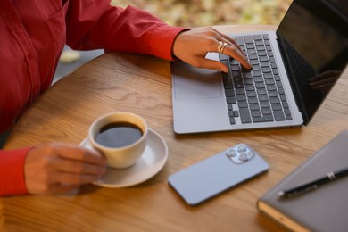 Photo of Woman with cup of coffee working on laptop at wooden table in cafe, closeup