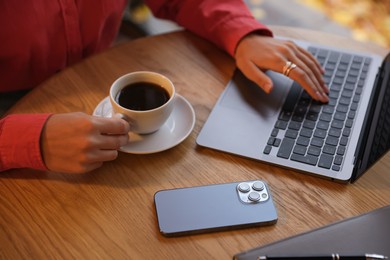 Photo of Woman with cup of coffee working on laptop at wooden table in cafe, closeup