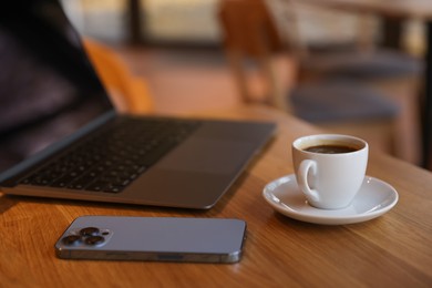 Photo of Laptop, cup of coffee and smartphone on wooden table in cafe, closeup