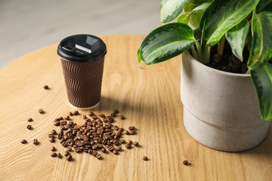 Photo of Paper cup, coffee beans and potted plant on wooden table indoors, closeup. Mockup for design