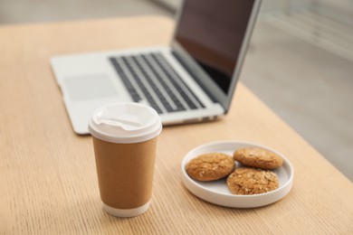 Photo of Paper cup, plate with cookies and laptop on wooden table indoors. Mockup for design