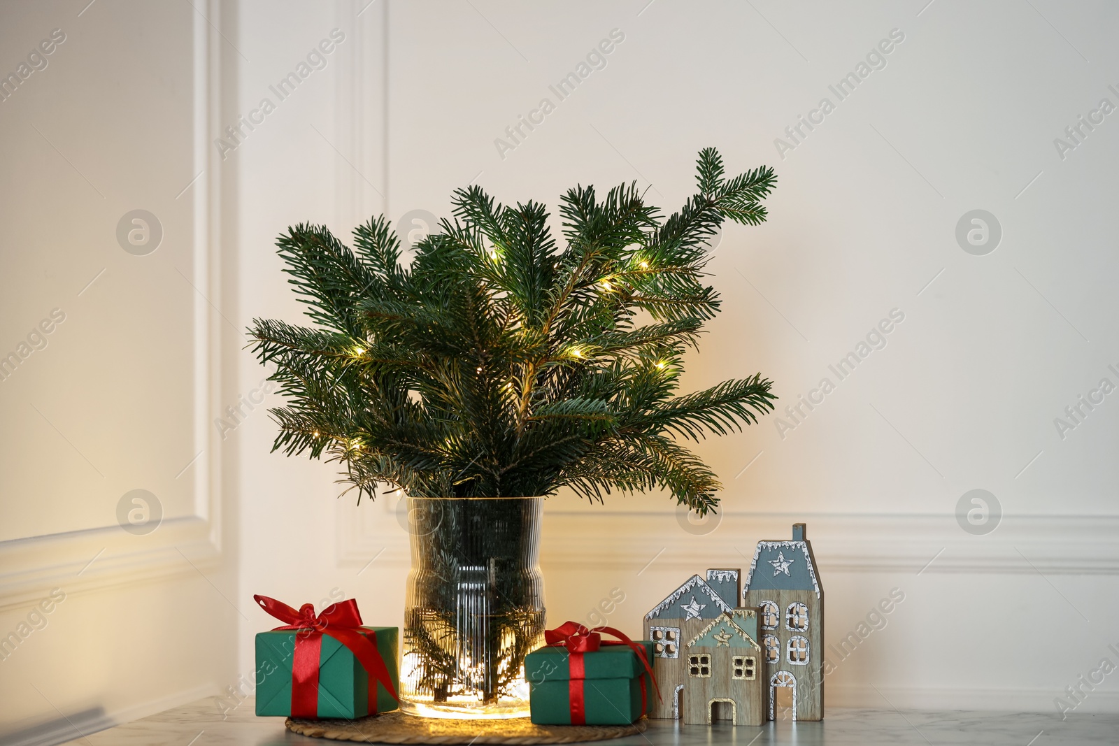 Photo of Beautiful fir tree branches decorated with festive lights and Christmas gifts on table near white wall