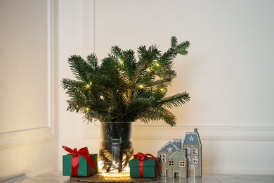 Photo of Beautiful fir tree branches decorated with festive lights and Christmas gifts on table near white wall