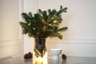 Photo of Beautiful fir tree branches decorated with Christmas lights and cones on table near white wall