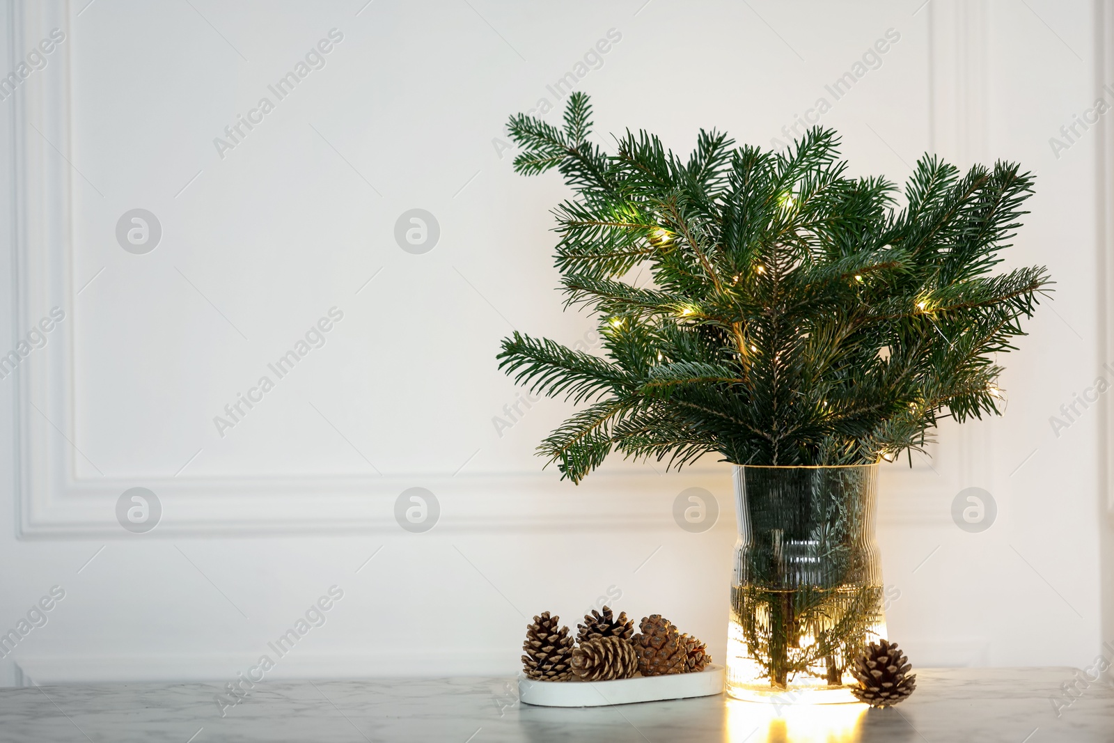 Photo of Beautiful fir tree branches decorated with Christmas lights and cones on table near white wall. Space for text