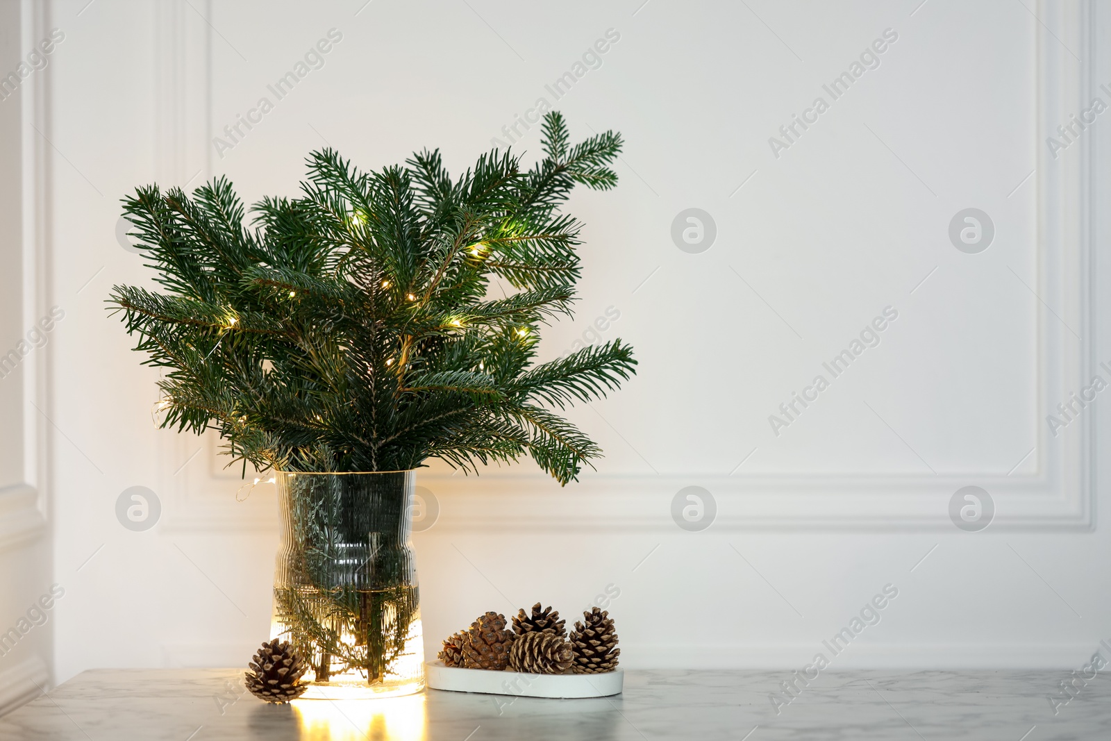 Photo of Beautiful fir tree branches decorated with Christmas lights and cones on table near white wall. Space for text