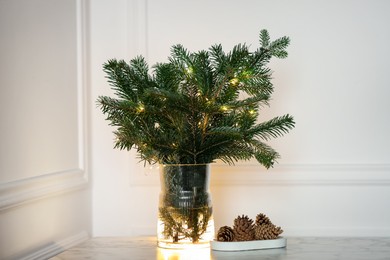 Photo of Beautiful fir tree branches decorated with Christmas lights and cones on table near white wall