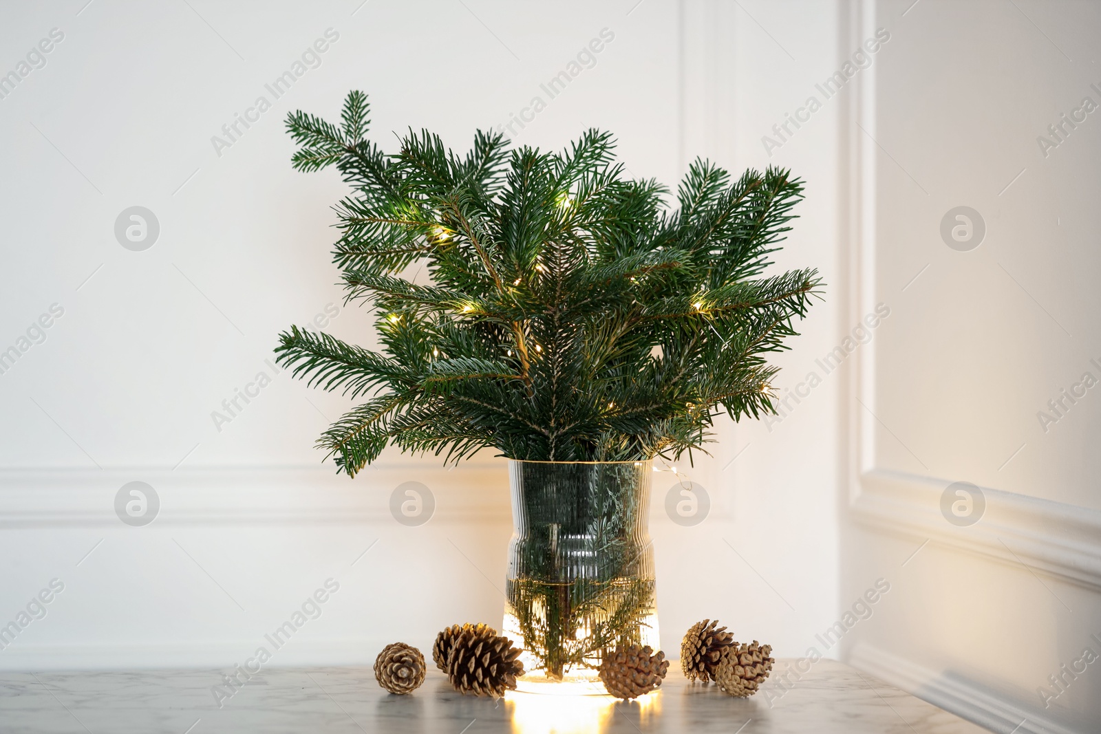 Photo of Beautiful fir tree branches decorated with Christmas lights and cones on table near white wall