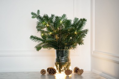 Photo of Beautiful fir tree branches decorated with Christmas lights and cones on table near white wall