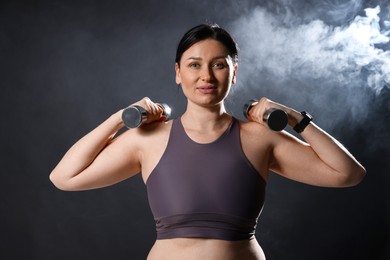 Photo of Plus size woman in gym clothes holding dumbbells on dark background with smoke