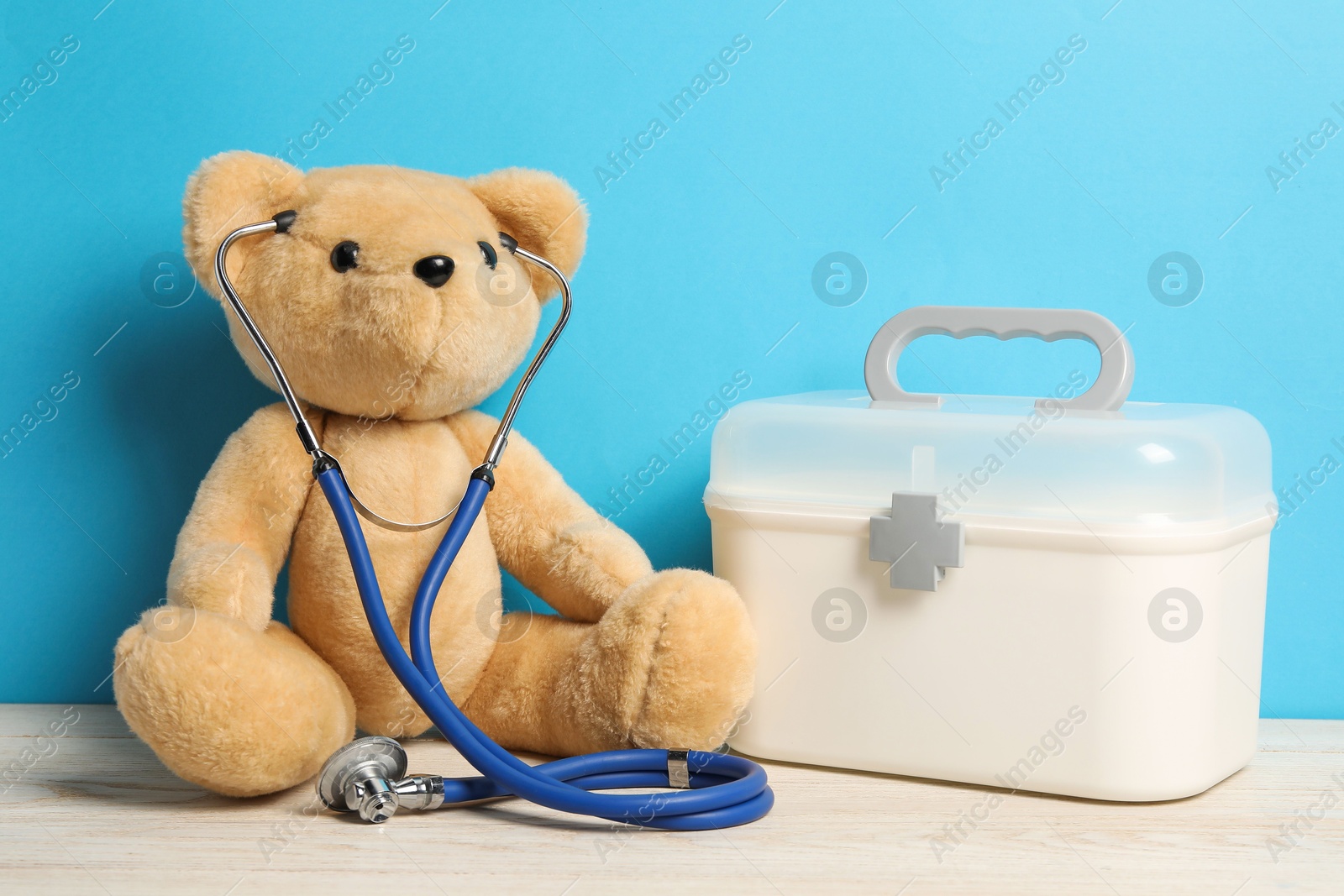 Photo of Pediatrics concept. Teddy bear with stethoscope and first aid kit on wooden table against light blue background